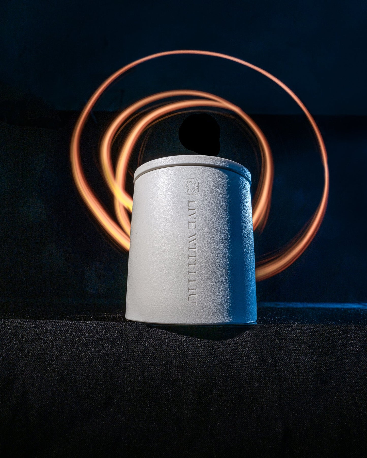 Essence candle in a ceramic jar, sitting on the edge of a table. The room is dark, but the yellow lightpainting in the background forms multiple circles to close the cycle with a grounded feel.
