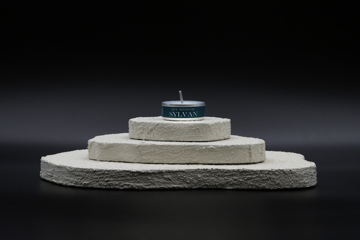 A shot of Sylvan in tealights, placed on top of a three-layered white flat rocks, with black background. Rain forest inspired refreshing scent.